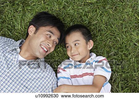 Stock Image - Father and son (7-9) lying on grass looking in eyes elevated view. Fotosearch