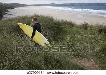 Picture - Man carrying surfboard down hill torwards sea back view. Fotosearch