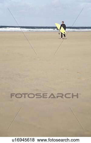 Stock Photo - Man carrying surfboard standing on beach back view. Fotosearch