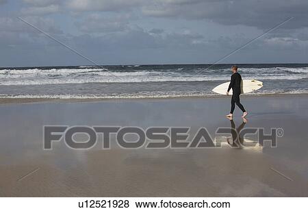 Stock Photo - Man carrying surfboard walking on beach. Fotosearch
