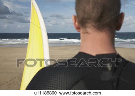 Stock Image - Man holding surfboard on beach looking at sea back view close up. Fotosearch