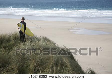 Stock Image - Man holding surfboard on cliff looking at sea back view. Fotosearch