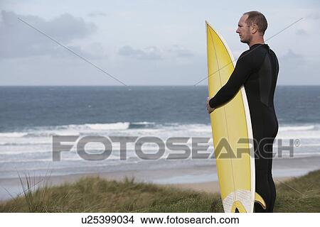 Picture - Man holding surfboard on hill looking at sea side view. Fotosearch