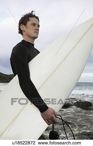 Stock Image - Man holding surfboard on rocky beach side view. Fotosearch
