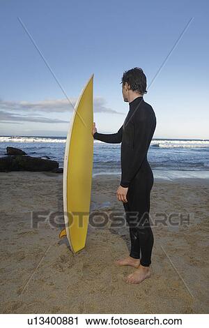 Stock Image - Man holding surfboard standing on beach side view. Fotosearch