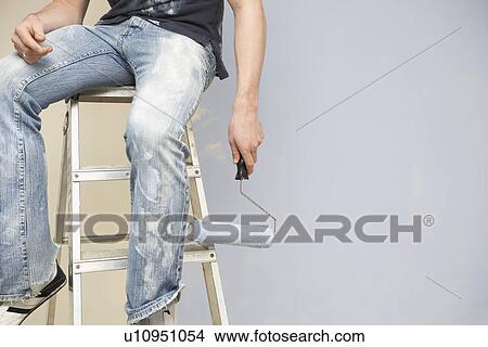 Picture - Man sitting on ladder holding paint roller low section. Fotosearch