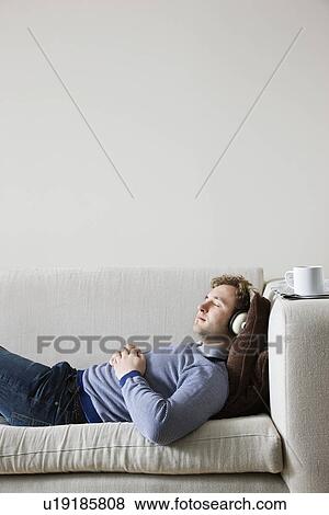 Stock Photo - Man wearing headphones lying on sofa in living room side view. Fotosearch