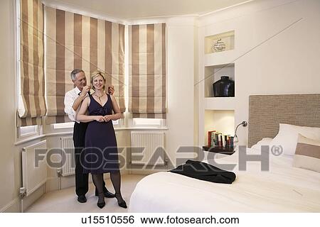 Stock Photograph - Middle-aged couple in bedroom man helping woman with necklace. Fotosearch