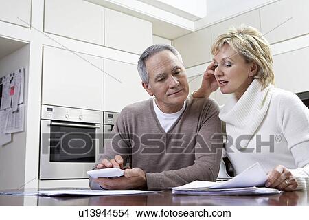 Middle-aged couple sitting in kitchen counting bills using calculator View Large Photo Image Picture - Middle-aged couple sitting in kitchen counting bills using calculator. Fotosearch