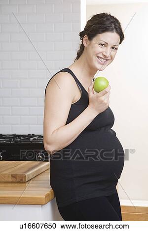 Stock Image - Pregnant woman eating apple in kitchen portrait. Fotosearch
