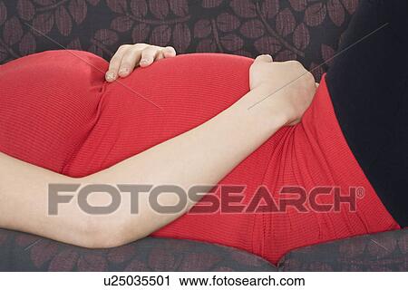 Stock Image - Pregnant woman relaxing on sofa mid section. Fotosearch