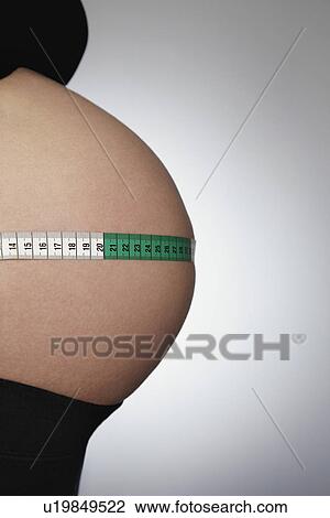 Stock Image - Pregnant woman with tape measure around abdomen mid section side view studio shot. Fotosearch
