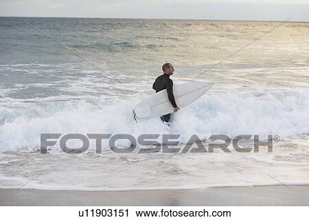 Stock Image - Surfer carrying surfboard in sea side view. Fotosearch