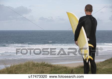Stock Image - Surfer holidng surfboard on hill looking at sea back view. Fotosearch