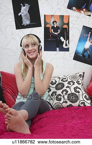 Stock Image - Teenage girl (16-17) sitting on bed listening to music. Fotosearch