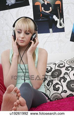 Stock Image - Teenage girl (16-17) sitting on bed listening to music. Fotosearch