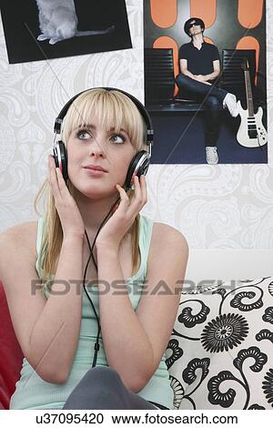 Stock Image - Teenage girl (16-17) sitting on bed listening to music. Fotosearch