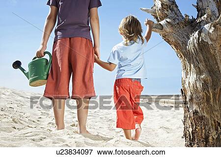 Stock Photo - Two boys (7-12) with watering can standing by dead tree on beach. Fotosearch