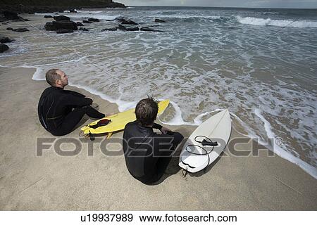 Two surfers sitting on beach looking at sea elevated view View Large Photo Image Stock Photo - Two surfers sitting on beach looking at sea elevated view. Fotosearch