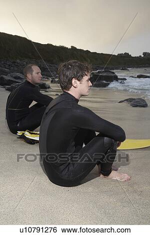 Stock Photograph - Two surfers sitting on beach looking at sea side view. Fotosearch