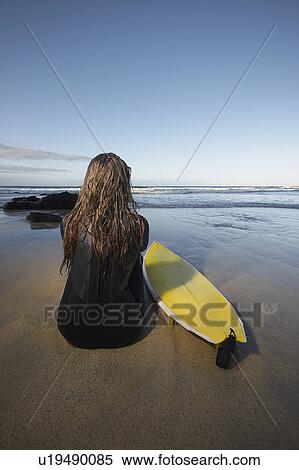 Stock Photography - Woman sitting by surfboard on beach back view. Fotosearch