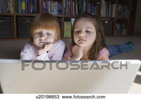 Stock Image - Young girls (5-6) lying on floor using laptop contemplating with hand on chin. Fotosearch
