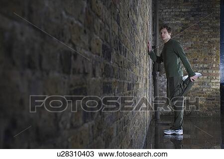 Stock Image - Young man doing leg stretch against brick wall. Fotosearch