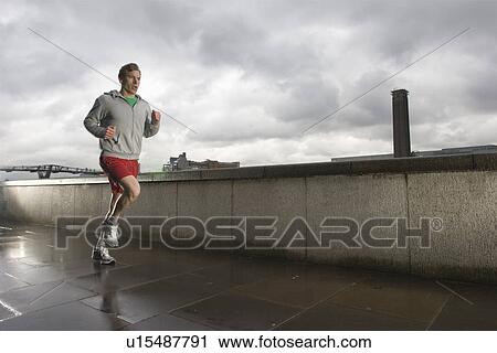 Stock Image - Young man jogging besides the Thames in London on stormy day. Fotosearch