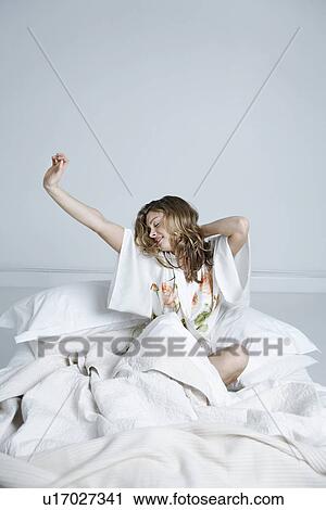 Stock Image - Young woman in dressing gown stretching in bed. Fotosearch