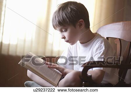 Stock Image - Boy sits cross-legged on chair reading book. Fotosearch
