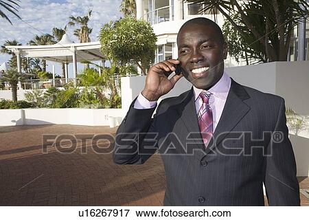 Stock Photo - Businessman stands holding mobile phone in front of hotel. Fotosearch