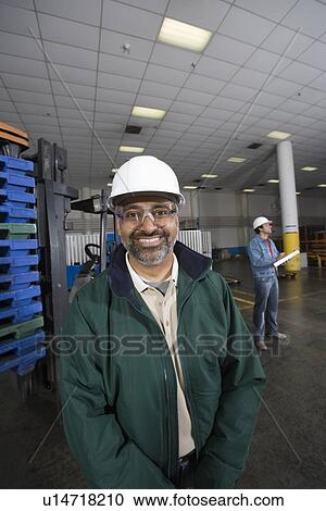 Stock Image - Cheerful man standing in factory wearing hart hat. Fotosearch