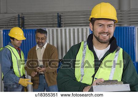 Stock Image - Cheerful man standing in factory colleagues in background. Fotosearch