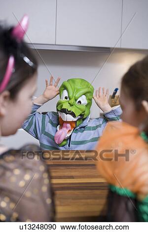 Stock Image - Child wearing mask scaring two girls in kitchen. Fotosearch