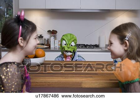 Stock Photograph - Child wearing mask scaring two girls in kitchen. Fotosearch