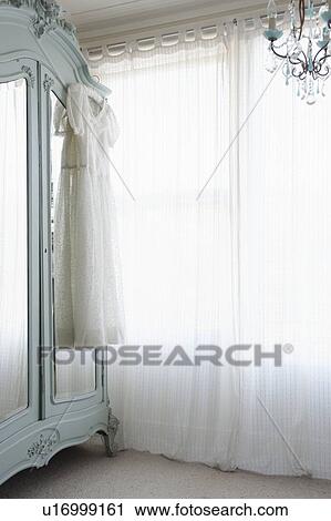 Stock Image - Christening gown on wardrobe at window with net curtains. Fotosearch