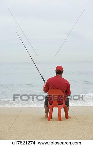 Picture - Man in red tshirt fishing from red plastic chair Cape Town South Africa. Fotosearch