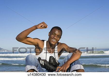 Stock Image - Man sitting on beach with with football boots round his neck cheering. Fotosearch