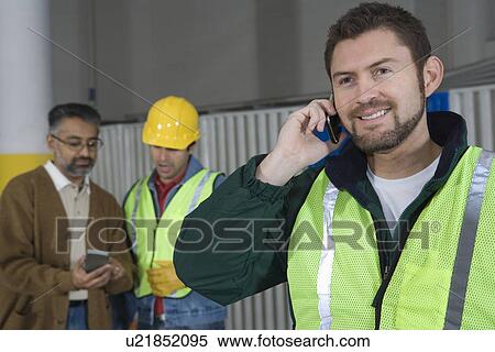 Stock Photography - Man standing in factory using telephone colleagues in background. Fotosearch