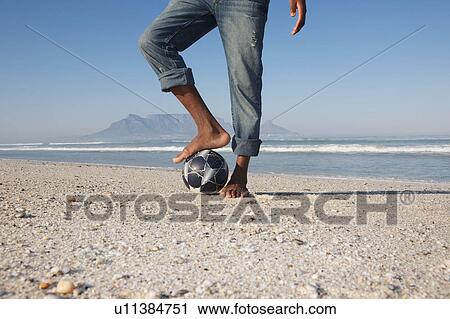Stock Image - Mans foot on soccer ball beach scene. Fotosearch