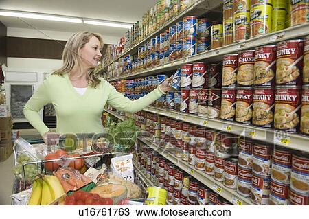 Stock Image - Mature woman selects tinned goods in supermarket aisle. Fotosearch