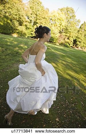 Stock Image - Mid-adult bride running in park. Fotosearch