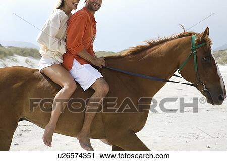 Stock Image - Mid-adult couple riding horse on beach. Fotosearch