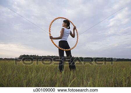 Stock Image - Mid adult woman stands with hula hoop in a field. Fotosearch