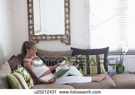 Stock Image - Pregnant woman reclining on sofa with book. Fotosearch