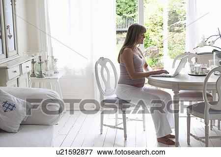 Stock Photo - Pregnant woman sitting at table using laptop. Fotosearch
