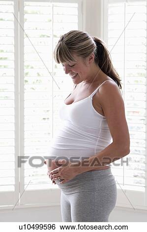 Stock Photograph - Pregnant woman standing near window touching stomach. Fotosearch