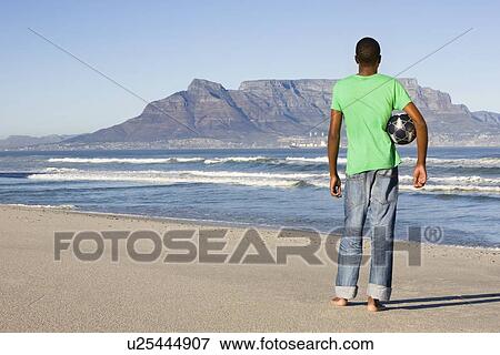 Rear view of man standing with football Table Mountain beach View Large Photo Image Stock Photo - Rear view of man standing with football Table Mountain beach. Fotosearch