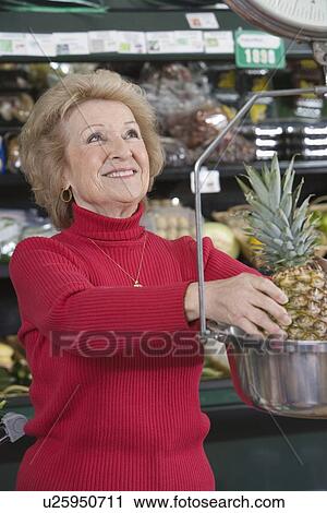 Stock Image - Senior woman weighing pineapple in supermarket. Fotosearch