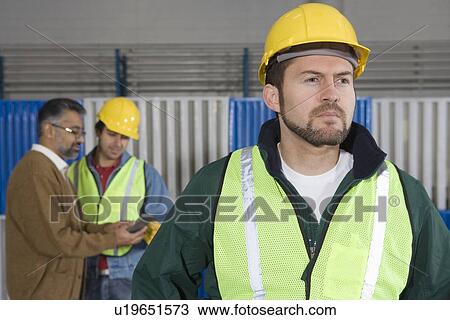 Stock Image - Serious man standing in factory colleagues in background. Fotosearch